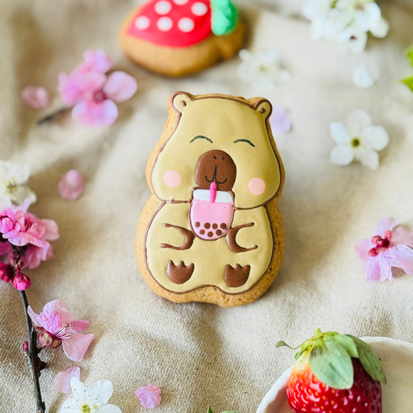 Decorative cookie shaped like a capybara with a cup, surrounded by strawberries and flowers.